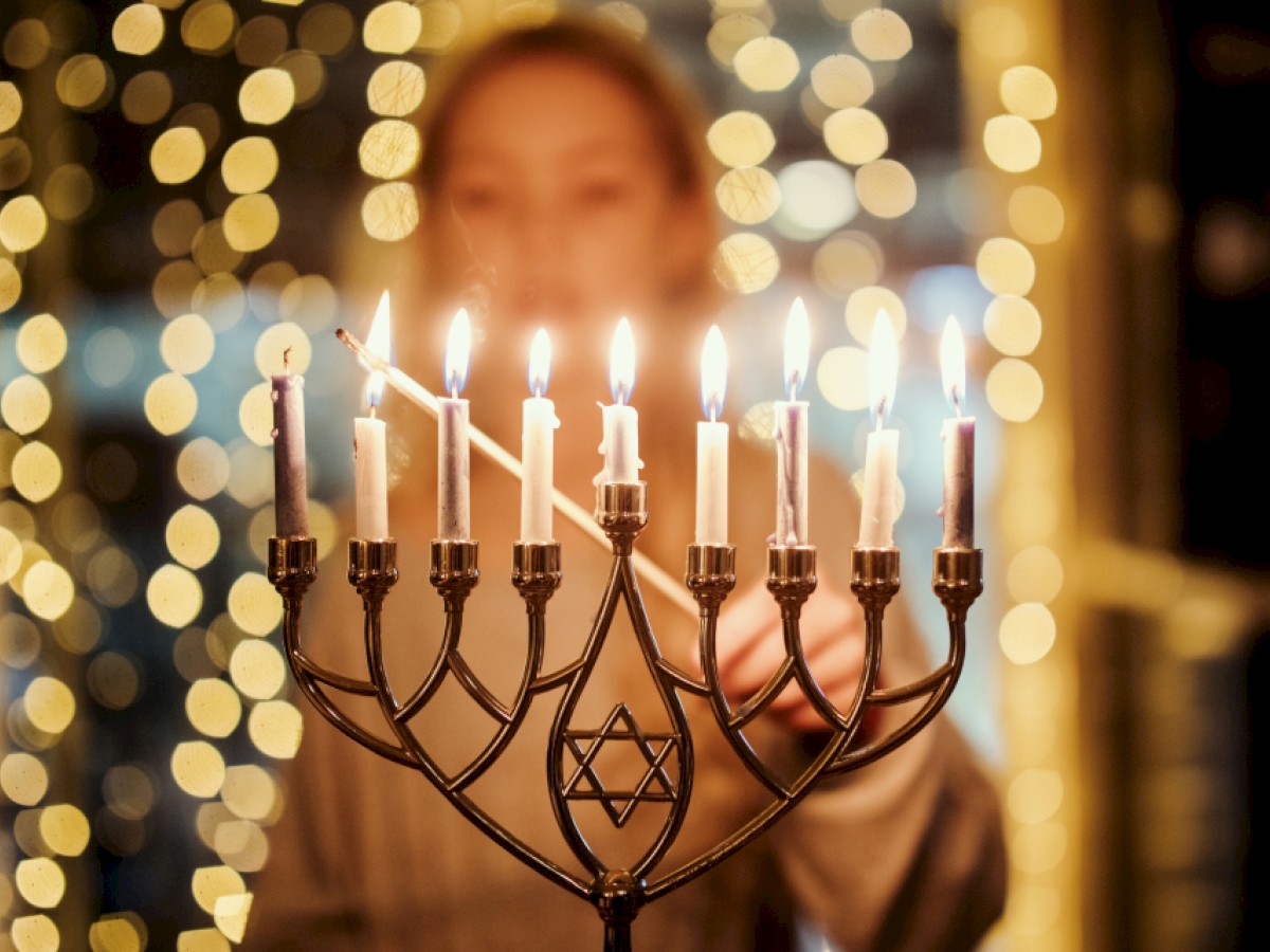 A person is lighting candles on a menorah with a blurred background of festive lights.