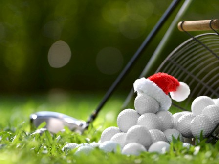 The image shows a pile of golf balls, with one wearing a small Santa hat, next to a tipped basket and a golf club on grass.