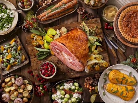 An assortment of holiday foods including roast ham, roasted vegetables, pies, fresh salads, carrot dishes, cranberry sauce, and garnishes on a table.