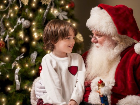 A child sitting on Santa's lap next to a decorated Christmas tree, both smiling and looking at each other warmly under festive lights.