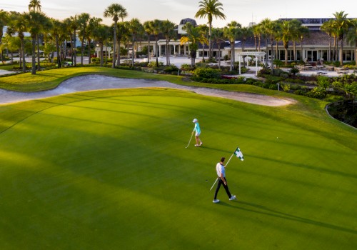 Two people are walking on a golf course with a clubhouse and palm trees in the background.