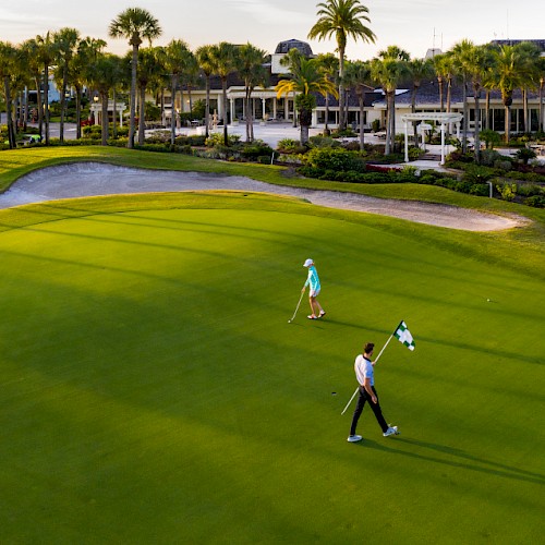 Two people are walking on a golf course with a clubhouse and palm trees in the background.