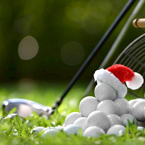 A pile of golf balls, one with a Santa hat, next to a basket and golf club on grass.