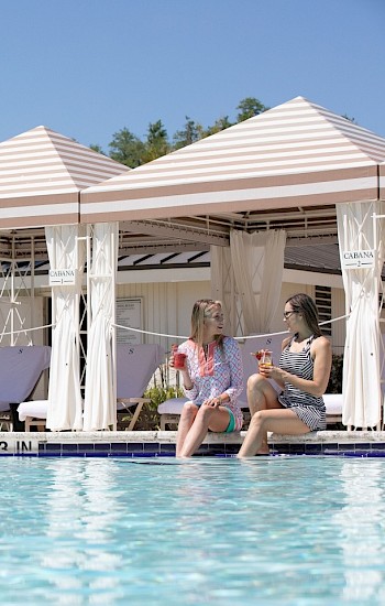 Two people sitting by a pool with drinks, under striped cabanas and palm trees in the background, enjoying a sunny day.