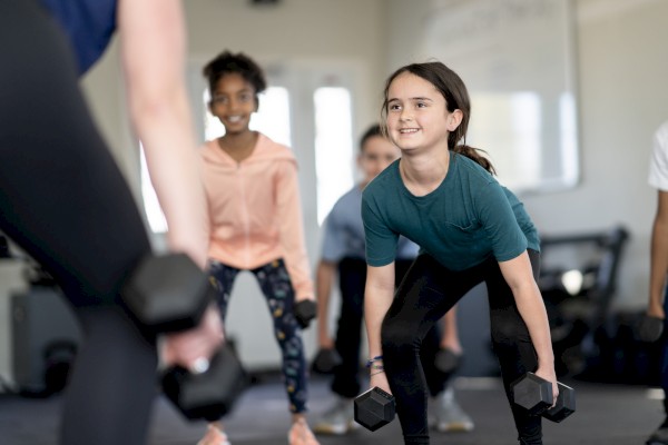 Children are participating in a fitness class, lifting dumbbells while following an instructor's guidance.