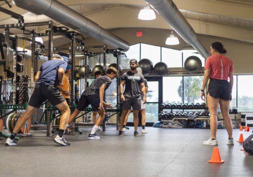 A group of people in a gym engage in exercise and training activities near weightlifting equipment and traffic cones, under a high ceiling.