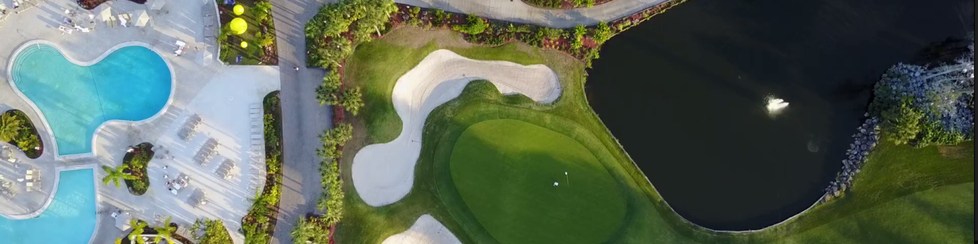 This aerial image shows a golf course with sand traps, a putting green, a pond, and nearby swimming pools surrounded by landscaping.