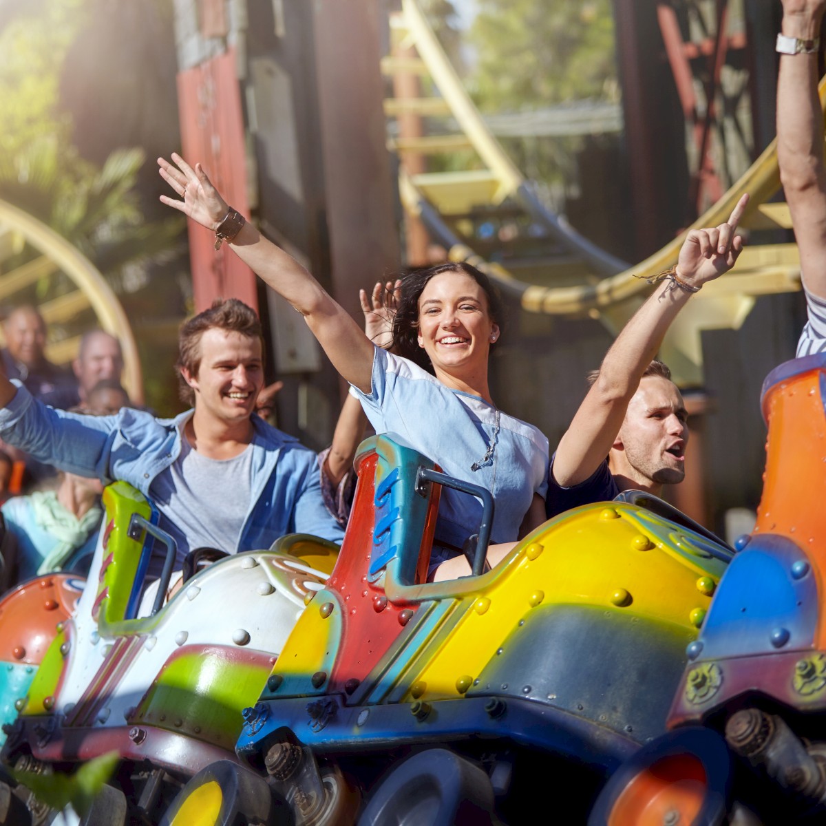 People are enjoying a colorful roller coaster ride, with hands raised and smiles, against a backdrop of amusement park scenery.