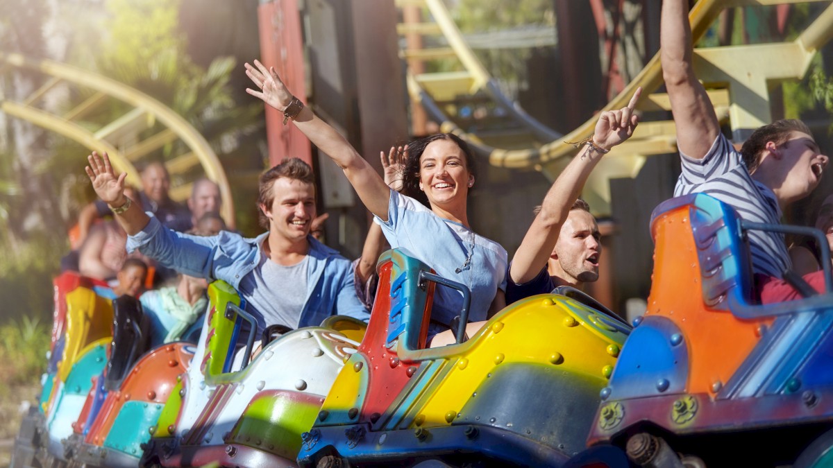 People are enjoying a colorful roller coaster ride, with hands raised and smiles, against a backdrop of amusement park scenery.