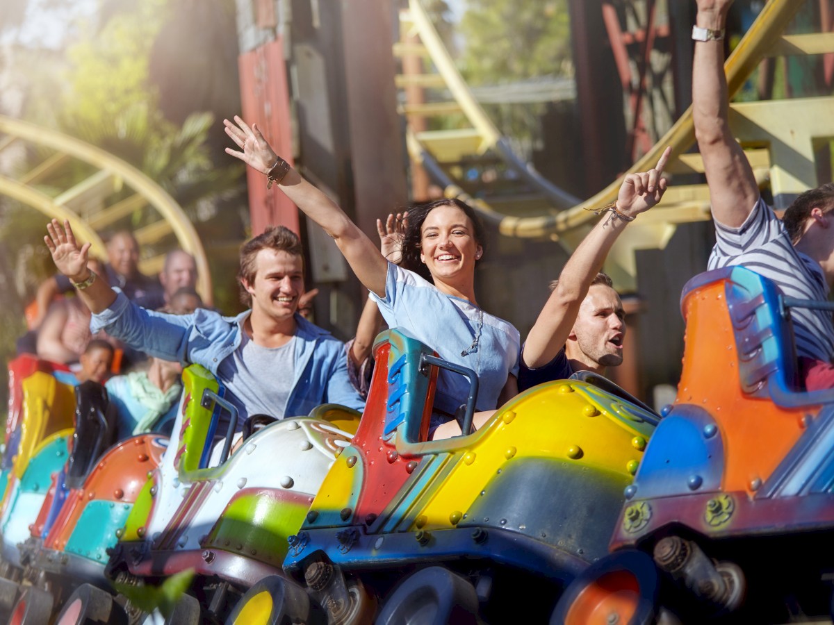 People are enjoying a colorful roller coaster ride, with hands raised and smiles, against a backdrop of amusement park scenery.