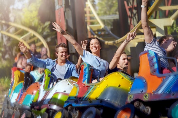 People enjoying a colorful roller coaster ride, smiling and raising their hands, with tracks and greenery visible in the background.