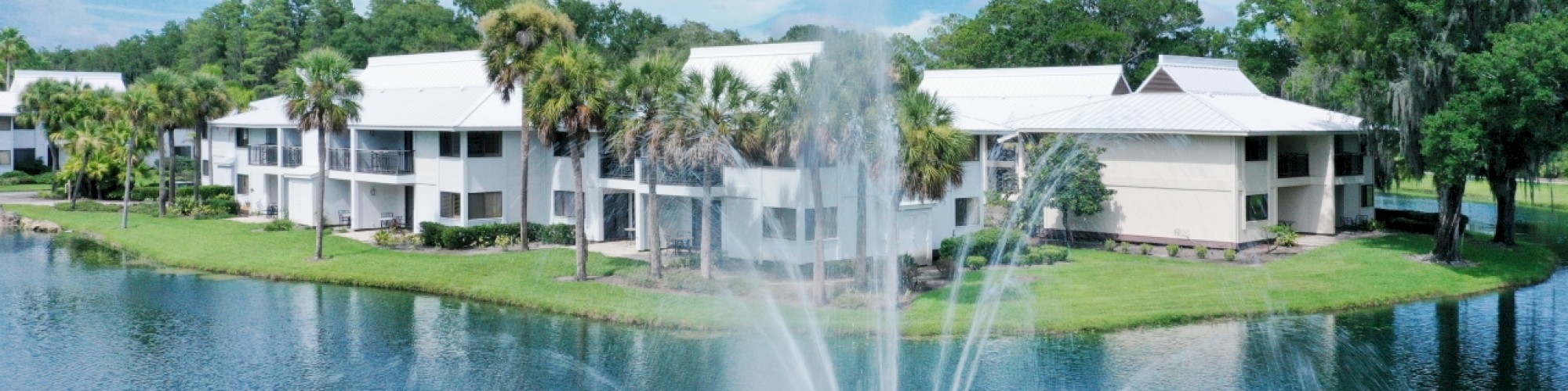 A scenic view of white-roofed buildings by a lake with a fountain and lush greenery under a clear blue sky.