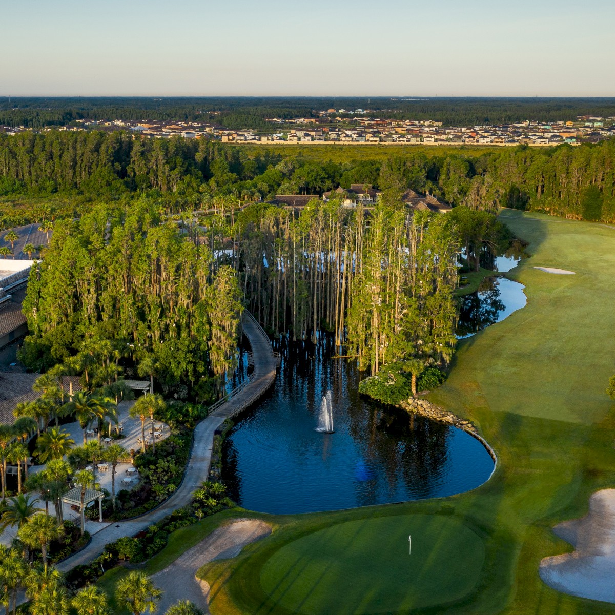 Aerial view of a golf course with sand traps, adjacent buildings, a water feature with trees, and expansive green areas under a blue sky.