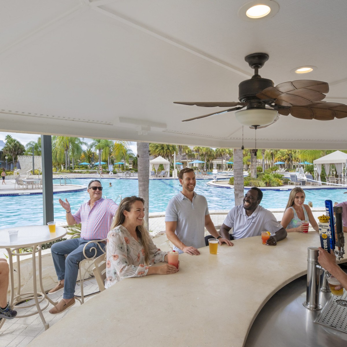 People sitting at a poolside bar enjoying drinks, with a bartender serving them. A fan is on the ceiling above the counter.