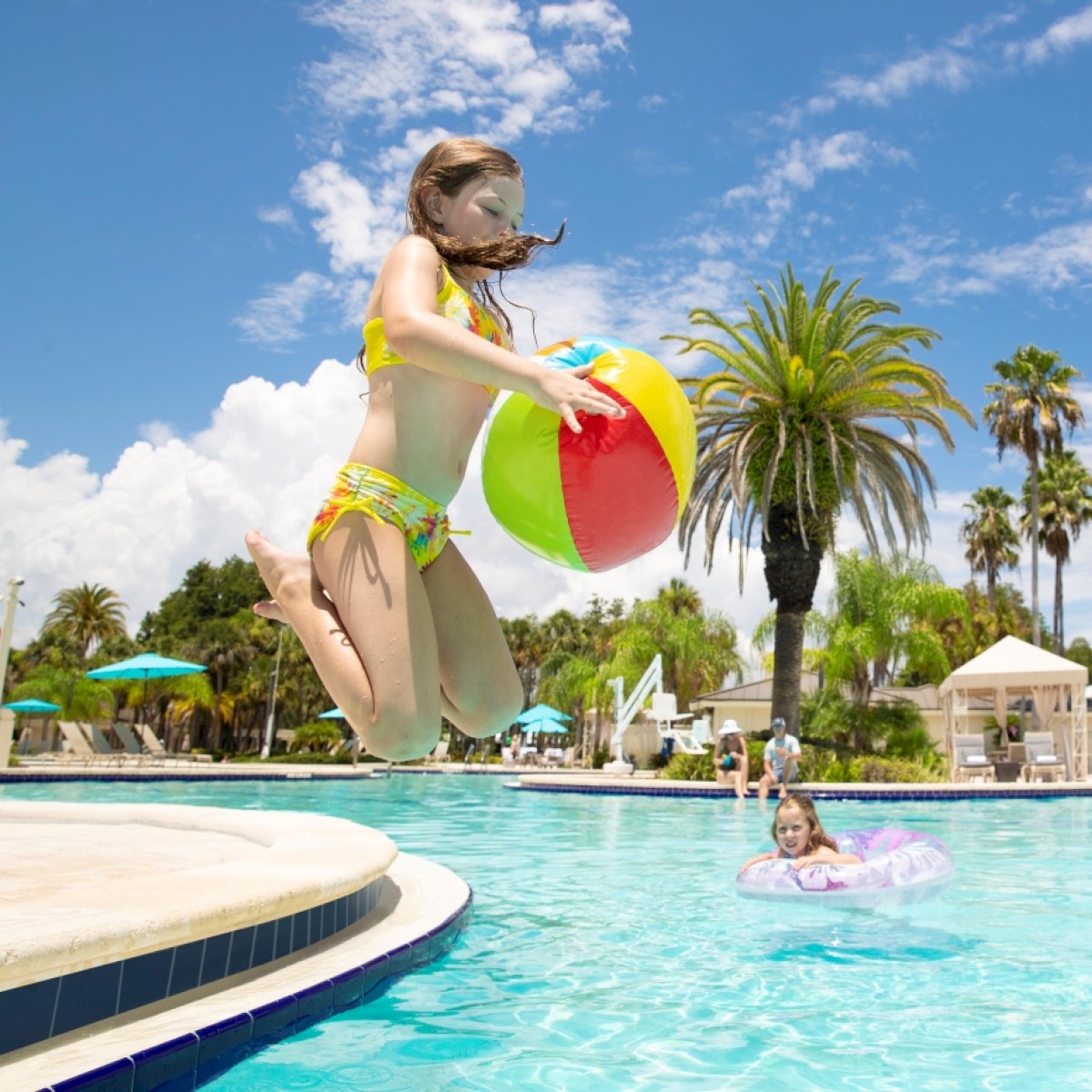 A child jumps into a pool with a beach ball while another floats on an inflatable ring; palm trees and blue sky in the background.