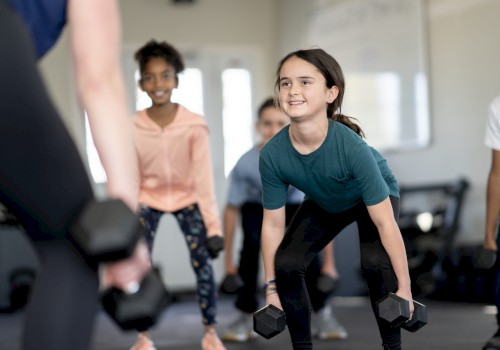 A group of kids is in a fitness class, lifting dumbbells, led by an instructor. Everyone appears engaged and smiling.