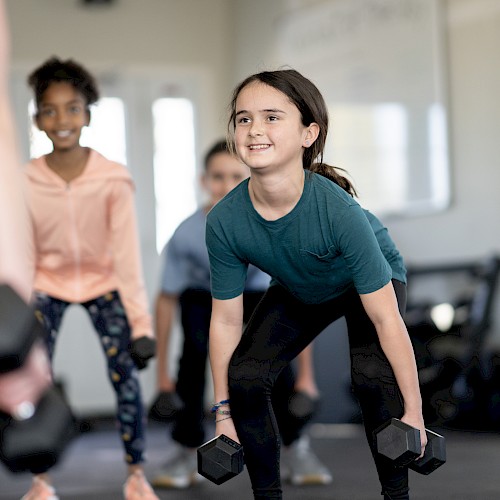 A group of kids is in a fitness class, lifting dumbbells, led by an instructor. Everyone appears engaged and smiling.