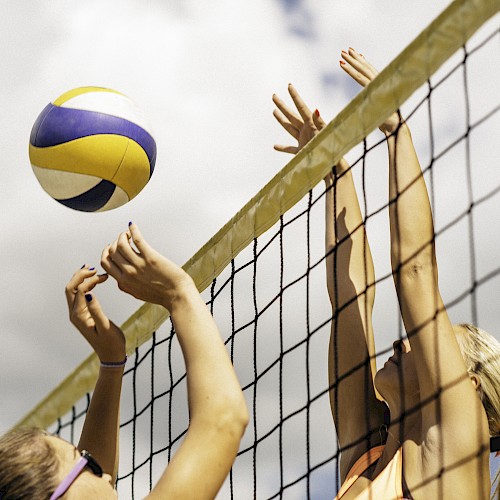 Two players at a volleyball net, one attempting to block a ball while the other prepares to spike it.