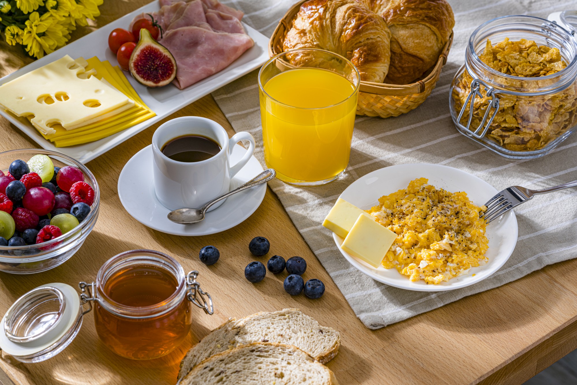 A breakfast spread with scrambled eggs, cheese slices, figs, ham, croissants, cereal, coffee, juice, honey, fruit salad, and bread.