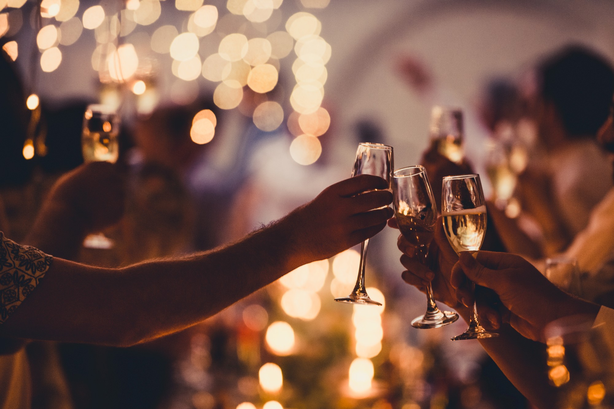 People clinking champagne glasses in a celebratory setting with warm, golden lights in the background, creating a festive atmosphere.