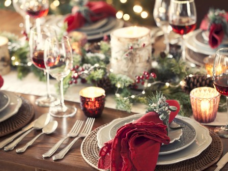 A festive table setup with candles, red napkins, wine glasses, and greenery. Plates and cutlery are elegantly arranged for a holiday meal.