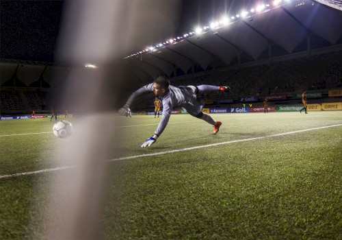 A soccer goalkeeper is diving to stop a ball near the goal line during a match under stadium lights.