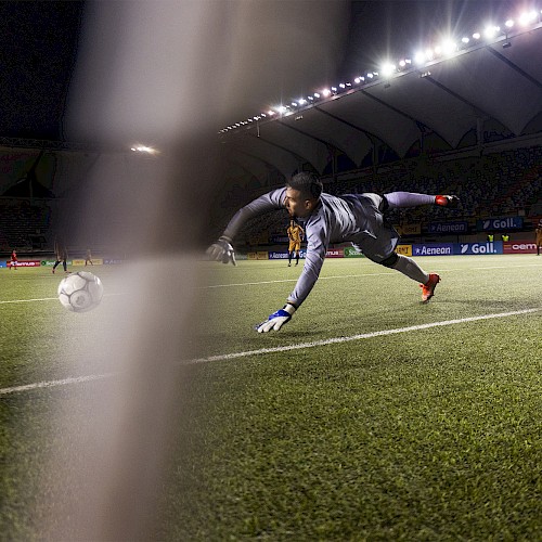 A soccer goalkeeper is diving to stop a ball near the goal line during a match under stadium lights.