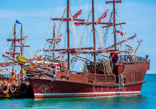 A large, ornate wooden sailing ship with multiple masts and red sails is docked at a harbor under a clear blue sky.