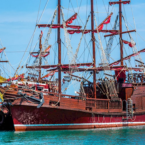 A large, ornate wooden sailing ship with multiple masts and red sails is docked at a harbor under a clear blue sky.