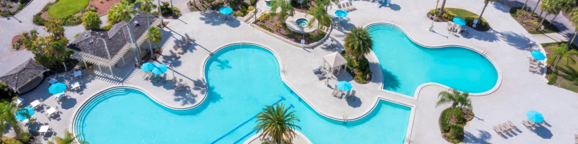 An aerial view of a resort swimming pool area with surrounding lounge chairs, umbrellas, and palm trees, near white-roofed buildings and pathways.