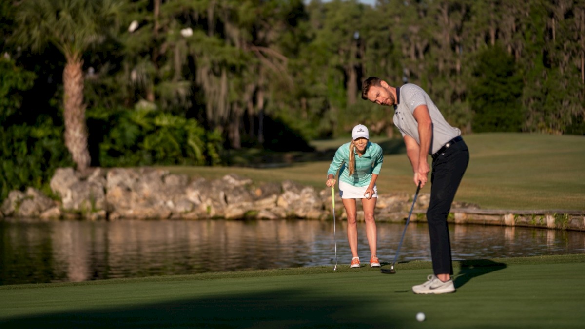 Two people are golfing on a green course, one putting while the other watches. They're surrounded by trees and a water hazard.