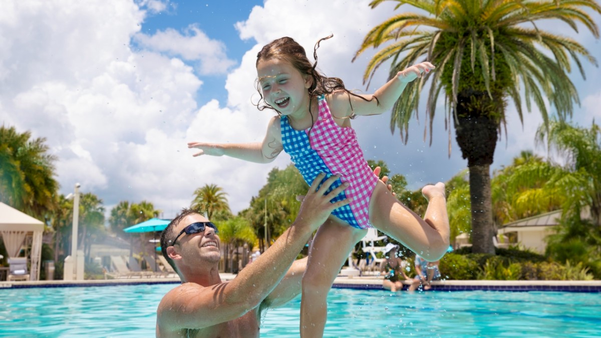 A man lifts a laughing child in a swimming pool, surrounded by palm trees under a sunny blue sky with clouds.