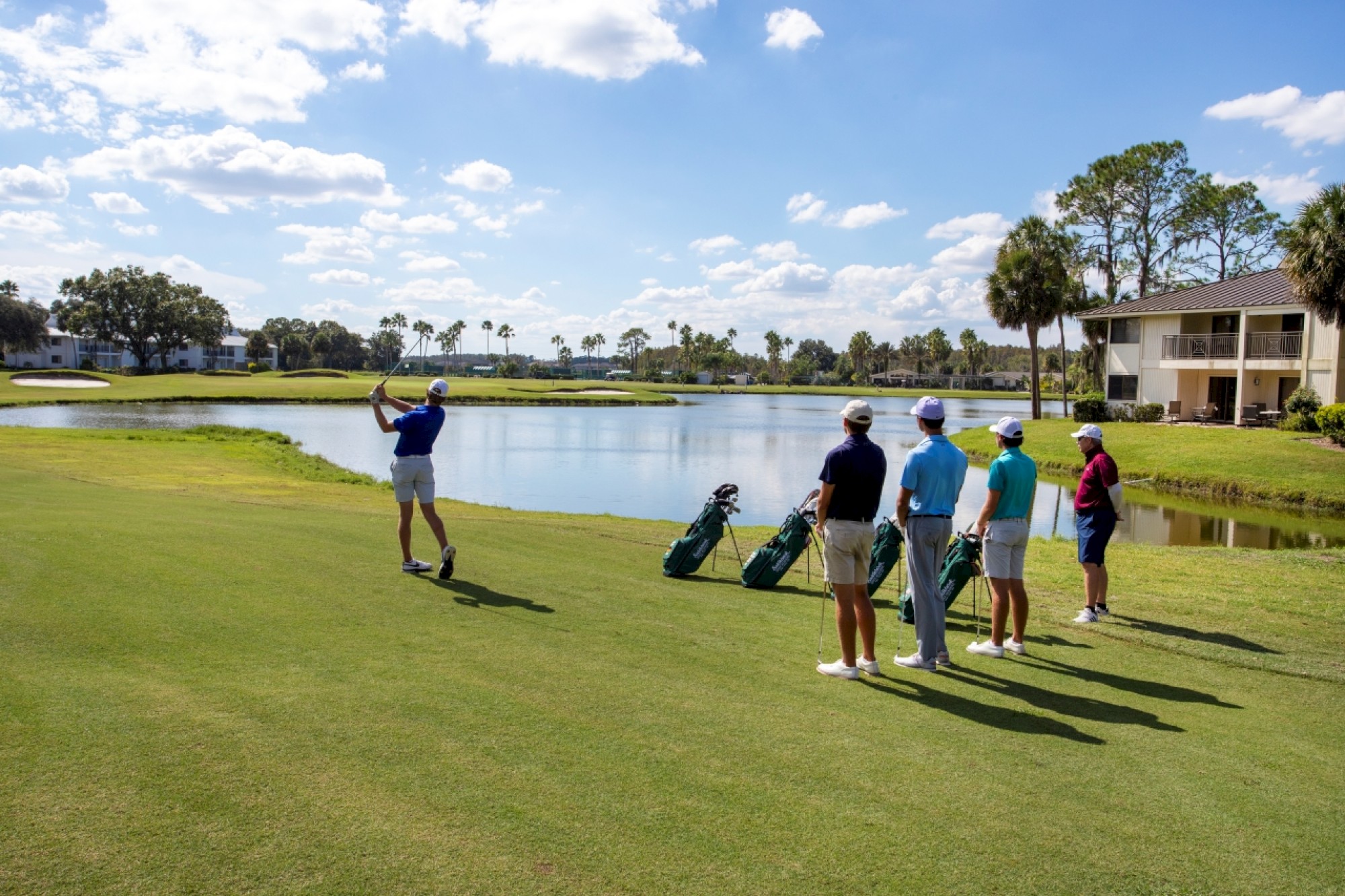 A group of golfers on a sunny course beside a water feature, with one person swinging a club while the others observe near golf bags.