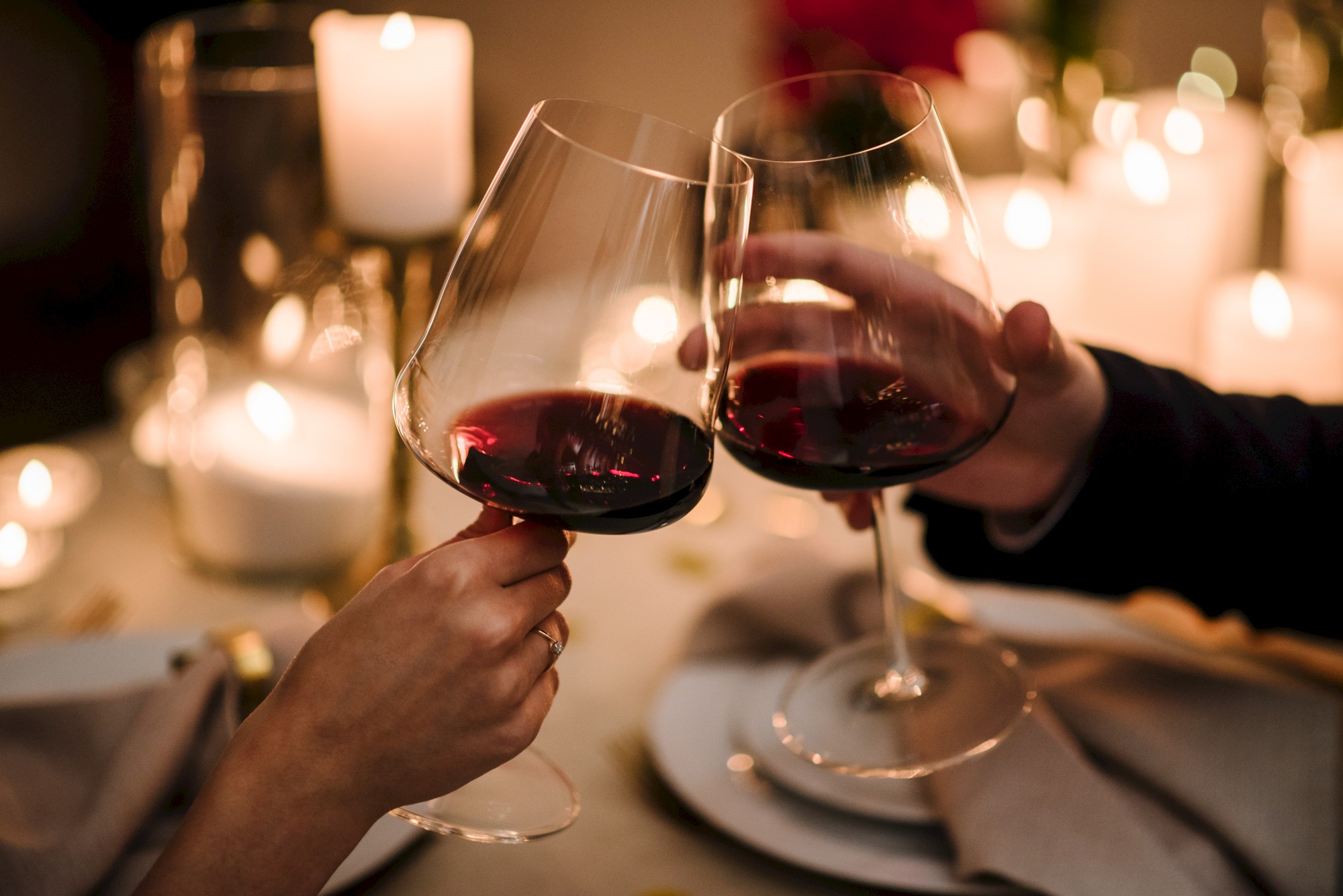 Two hands holding wine glasses in a toast with a romantic candlelit setting in the background.