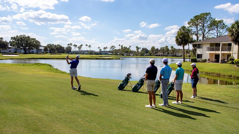 People on a golf course with clubs, near a pond, under a sunny sky