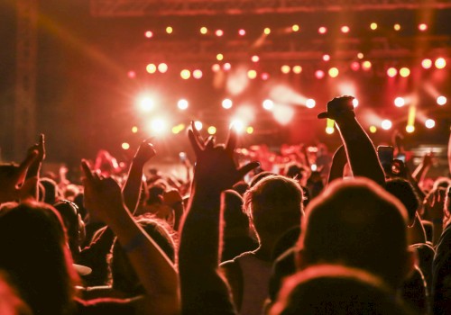 A crowd at a concert with hands raised, illuminated by bright stage lights in the background, creating a vibrant and energetic atmosphere.