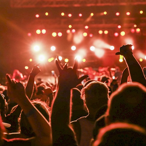 A crowd at a concert with hands raised, illuminated by bright stage lights in the background, creating a vibrant and energetic atmosphere.