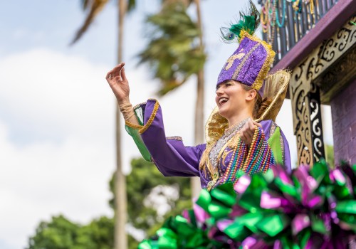 A person in vibrant, festive attire is cheerfully waving from a decorated float, set against a backdrop of palm trees and a bright sky.