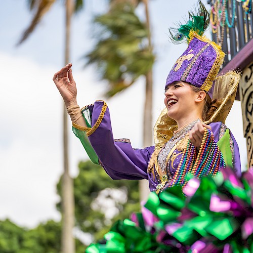 A person in vibrant, festive attire is cheerfully waving from a decorated float, set against a backdrop of palm trees and a bright sky.