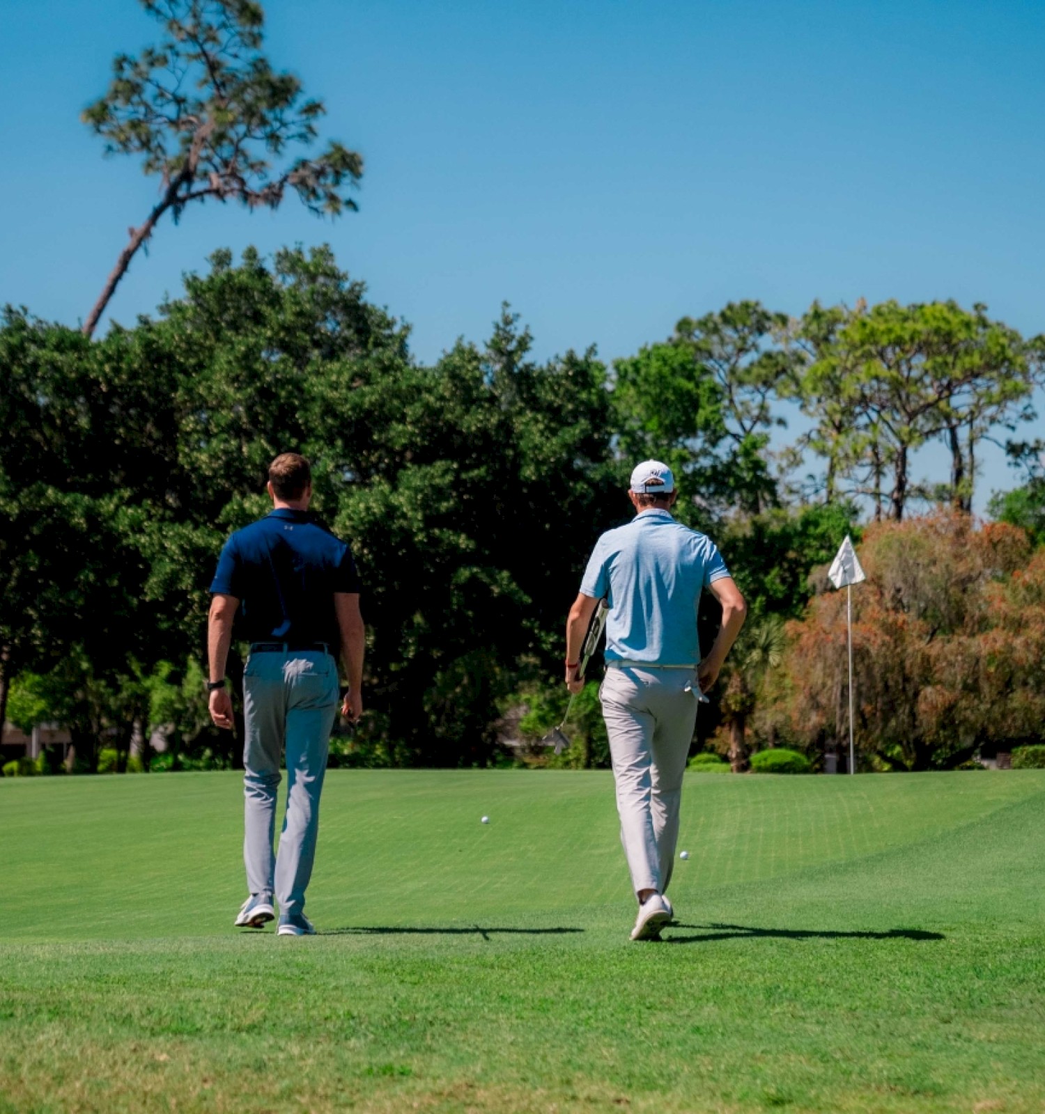 Two people walking on a golf course towards a flag, surrounded by greenery and trees under a clear blue sky.