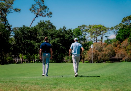 Two people walking on a golf course towards a putting green, with trees and a clear blue sky in the background.