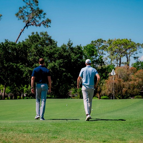 Two people walking on a golf course towards a putting green, with trees and a clear blue sky in the background.