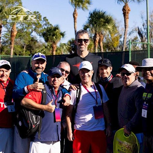 A group of people in sports attire posing outdoors with palm trees in the background, some wearing caps and sunglasses.