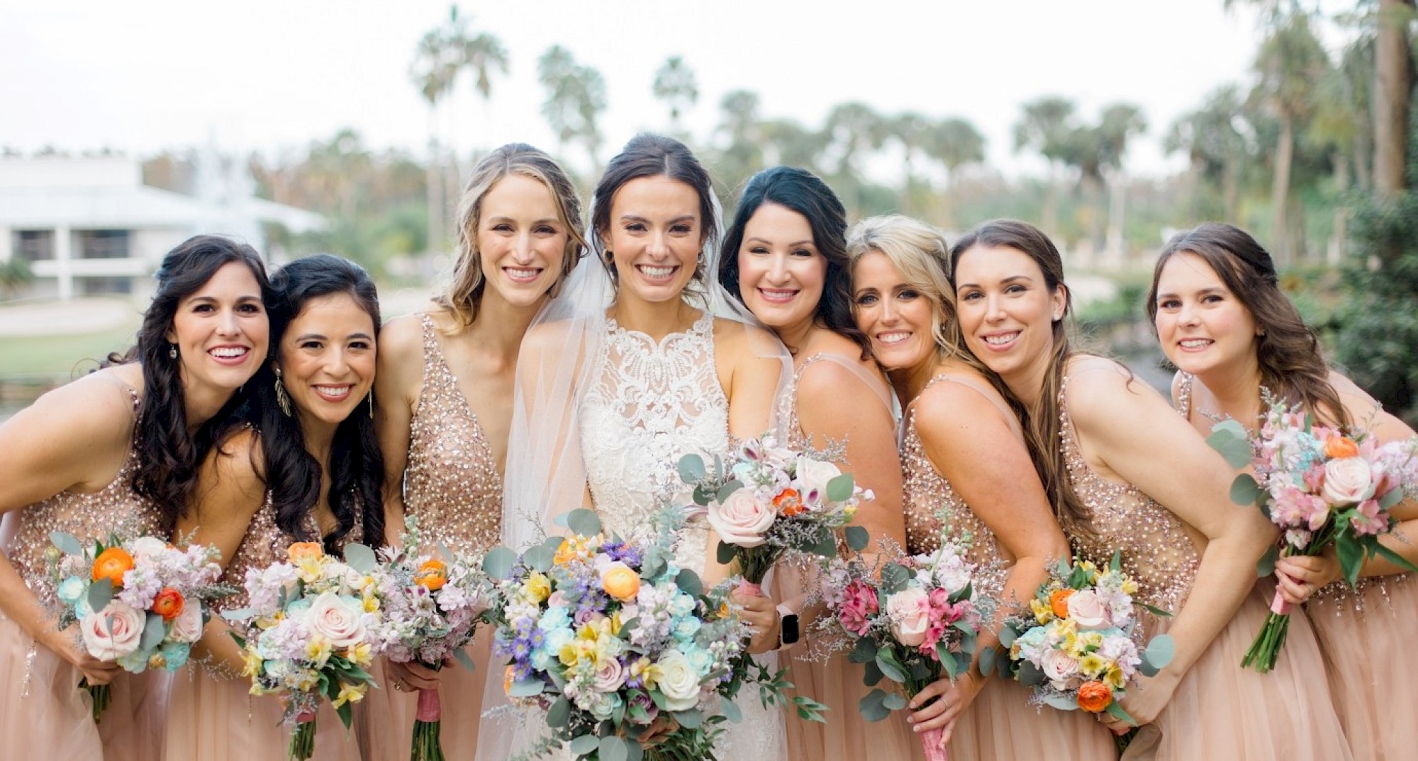 A bride and seven bridesmaids in matching dresses hold colorful bouquets, smiling outdoors with palm trees in the background.