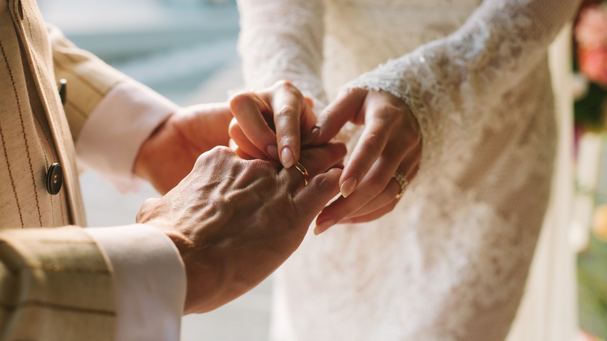 A bride is placing a ring on a groom's finger during a wedding ceremony, both in formal attire.