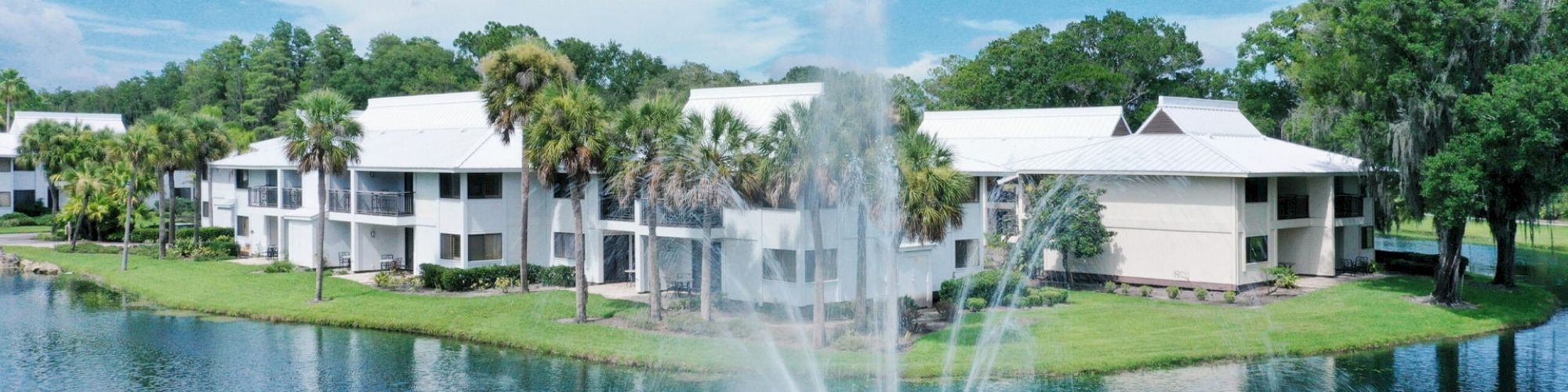 The image shows a serene lakeside view with a large fountain, surrounding green trees, and white residential buildings in the background.