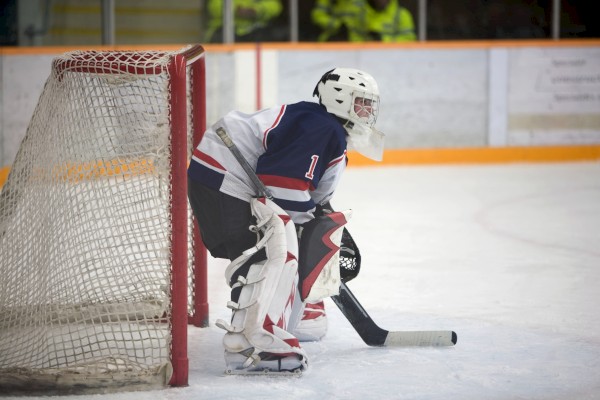 A hockey goalie in gear stands by the net on the ice, poised and ready in an arena setting.