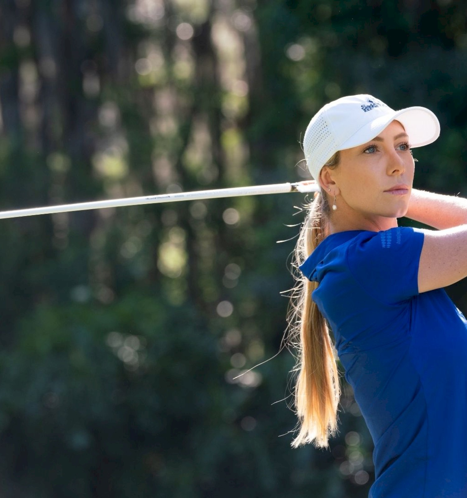 A golfer in a blue shirt and white cap is swinging a golf club outdoors, with trees in the background.