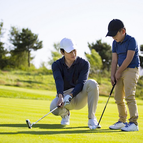 Two people on a golf course; one kneels, instructing the other on putting. Blue flag in the background, sunny day.