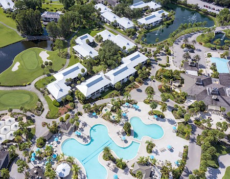 Aerial view of a resort with multiple pools, palm trees, landscaped gardens, and nearby golf course.